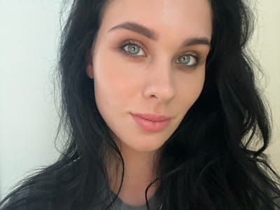 A close-up portrait depicts an adult female with long, dark, wavy hair and light-colored eyes, facing directly towards the viewer with a slight, neutral expression and subtly parted lips. She exhibits defined eyebrows, light eye makeup, and fair skin. A segment of a grey garment is visible at the bottom edge of the frame. The background is an unadorned, plain light-colored wall, indicating an indoor setting. The scene is located in 강서구, South Korea.