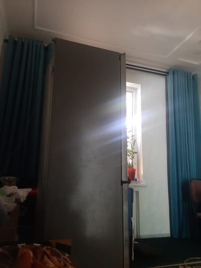 The image depicts the interior of a room, likely in Damburqut, Uzbekistan. A large, grey refrigerator is positioned in the foreground, partially obscuring a window. Sunlight streams through the window, creating lens flare. A potted plant sits on the windowsill. To the left and right of the refrigerator are teal-colored curtains. The ceiling has a decorative molding. In the lower left portion of the image, some blurred objects, possibly bags or containers, are visible. The floor appears to be carpeted in a dark color. The overall scene suggests a domestic setting with a focus on the refrigerator and the natural light entering the room.
