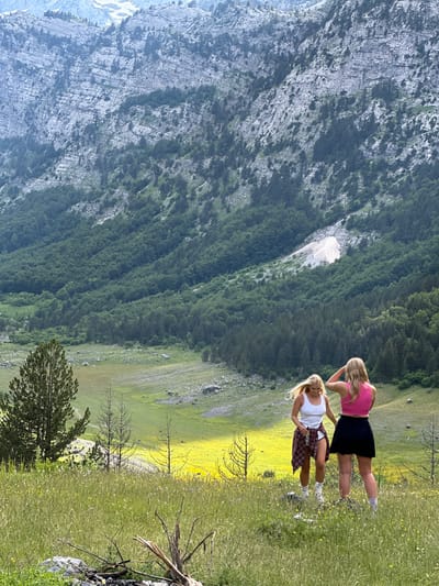 Two young women, with blonde hair, are standing in a grassy, flower-filled meadow. One woman wears a white tank top, red and black plaid shirt tied around her waist, white shorts, and white socks with sneakers. The other woman wears a pink crop top and a black skirt. They are facing away from the camera, looking towards a vast, green, forested mountain range. The mountain landscape is steep and rocky, with trees growing on its slopes. The sky is overcast. The location is identified as being in Shalë, Albania.