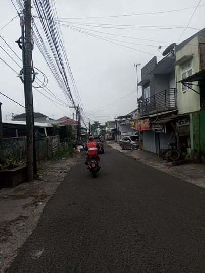 The image displays a street in the city of Houaythat, Laos, under an overcast, light grey sky. An individual on a dark motorcycle is centered in the mid-ground, traveling away from the camera. The rider wears a red and white long-sleeved shirt with "PELANG TENIS CLUB" printed on the back, and red shorts or pants. The motorcycle's rear license plate displays "2553 KB 09-24". The asphalt road surface exhibits minor wear.
On the left, a dark utility pole heavily laden with numerous black electrical and communication wires extends vertically. A concrete wall with visible graffiti and a green metal gate follows, adjacent to an area with sparse green vegetation and multiple black trash bags accumulated on the dirt and concrete shoulder. Various residential buildings with differing rooflines are visible further down this side.
On the right, a sequence of buildings lines a concrete sidewalk. The nearest structure features a light green lower facade and a grey upper level with a black metal balcony railing. A satellite dish and a television antenna are mounted on its upper exterior. Below a corrugated metal canopy, multiple signs for businesses are attached, including "TAMBAL BAN" (tire repair) and "KAISER". A discarded tire rim and what appears to be mechanical equipment are positioned near the "TAMBAL BAN" sign. Further down the street, a silver sedan is parked on the right, and other vehicles are visible. Palm trees are present in the distant background.