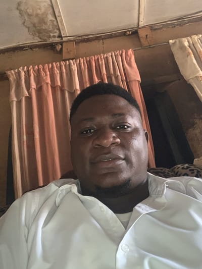 A medium shot, taken from a slightly high angle, shows a Black man from the chest up, looking directly at the camera. He has short, dark hair and a goatee. He is wearing a white button-down shirt, the collar of which is visible. The background appears to be indoors, with peach-colored curtains to the left and beige patterned curtains to the right. Above the curtains, the ceiling shows signs of water damage. A leopard-print fabric is visible to the right, behind the man's shoulder. The scene suggests a domestic setting, possibly in Tabu, Nigeria, given the context.