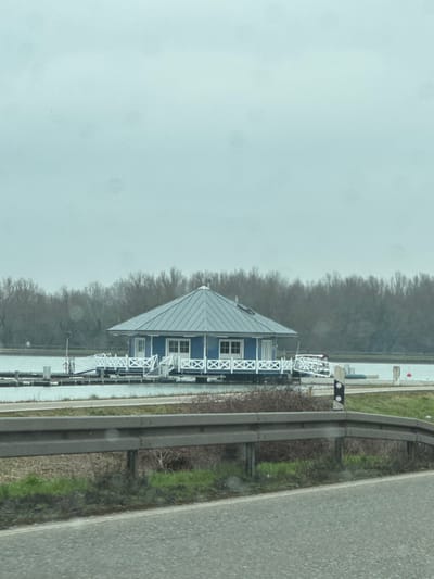 This image depicts a boathouse or floating structure situated on a body of water in Altenheim, Germany. The scene is observed from an elevated position, likely a roadside, under an overcast sky.
In the foreground, a dark asphalt road surface is visible, partially obscured by a grey metal guardrail. Below the guardrail, sparse green grass and brown vegetation grow. To the right of the guardrail, a white and black road marker post (Leitpfosten) stands.
The midground features a calm body of water. A prominent blue building, possibly octagonal or hexagonal in shape, rests on a white floating platform. The building has a light blue/grey conical roof with a small chimney or vent on top. Its exterior walls are blue with white trim around multiple windows and a white door on the visible side. White decorative railings with an "X" pattern surround the platform and extend horizontally across the water, connecting to other floating elements or docks.
In the background, a dense, horizontal line of bare deciduous trees defines the distant shoreline, extending across the entire width of the frame beneath the light grey sky. No individuals or active interactions are visible.