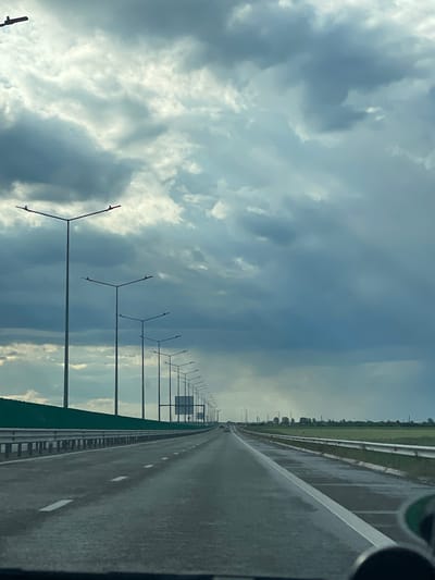 A slightly blurred image presents a view along a highway in Tunari, Romania. The perspective is from within a vehicle, as suggested by the dashboard and the angle of the shot. The highway is marked by a gray surface, delineated by white lane markers, and flanked by metal guardrails and a green barrier on either side. A series of uniformly spaced light poles line the left side of the road. The sky above is filled with dramatic, textured clouds in shades of gray and white. The image is oriented vertically.