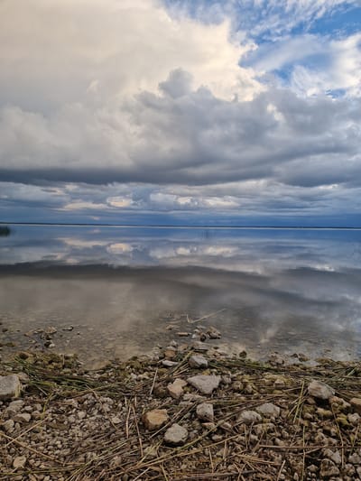 The image depicts a serene natural landscape with a calm body of water, likely a lake or the sea, under a dramatic, cloudy sky. The foreground is dominated by a stony shoreline covered with dry reeds and grass. The water's surface acts as a mirror, reflecting the textured clouds and the distant horizon, which appears as a thin blue line. Some sparse reeds or marsh grass are visible sticking out of the water near the left side. The overall impression is one of tranquility and a slightly overcast, possibly pre-storm atmosphere. The location is specified as the city of Īdeņa, Latvia.