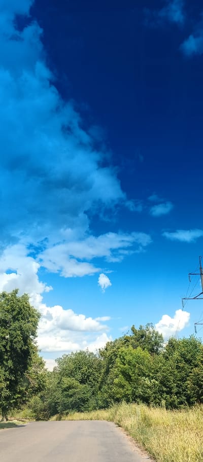 The image depicts a view of a paved road curving to the left, bordered by tall, dry grass and lush green trees. The sky is a vibrant, deep blue, with scattered white cumulus clouds. In the distance, to the right, a tall electrical pylon with insulators and wires is visible against the sky. The scene appears to be in a rural or semi-rural area, possibly on the outskirts of Kholodnianske, Ukraine, given the presence of the road, vegetation, and electrical infrastructure. There are no people or animals visible in the image.