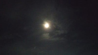 The image shows a night sky with the moon visible. The moon is bright and appears to be partially obscured by clouds. There are faint wisps of clouds around the moon, creating a hazy effect. The rest of the sky is dark and appears to be filled with a thin layer of clouds or haze. There is a faint green circular anomaly to the right of the moon. The image is grainy, indicating it was likely taken in low light conditions.