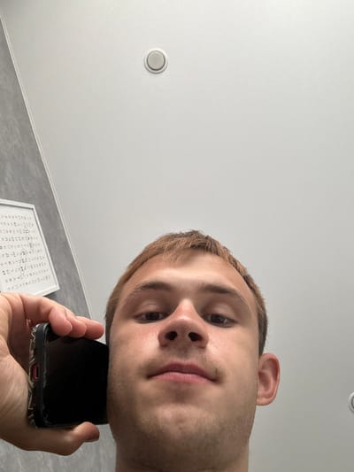 This media file is a selfie of a young man taken from a low angle, looking up at the camera. He has short, light brown hair and is holding a smartphone to his ear, as if on a call. His face is close to the phone, and his expression is neutral. The background appears to be a white ceiling with a circular light fixture. Below the ceiling, a gray textured wall is visible. In the lower right portion of the image, a framed piece of paper with what appears to be a word search puzzle is partially visible. The puzzle contains Cyrillic characters. The lighting suggests it is daytime. The location context provided is Tlustenkhabl, Russia.