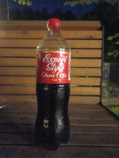 A medium shot captures a full, upright plastic bottle of "Export Style Classic Cola" against a backdrop of wooden planks. The bottle is mostly full, with the dark cola inside showing condensation droplets on the exterior, suggesting it's cold. The red cap is clearly visible at the top, and the label is predominantly red with white cursive text that reads "Export Style" and "Classic Cola." There's a small, stylized yellow star or emblem below the text. The bottle is positioned centrally on a wooden surface, possibly a bench or table, with the grain of the wood running horizontally. Behind the bottle, more wooden planks form a wall or barrier, also with horizontal grain. In the upper part of the image, blurred greenery and dark, indistinct shapes suggest foliage and trees, hinting at an outdoor setting. The lighting is dim, indicating it might be dusk or nighttime. The overall scene is still and quiet. No people are visible. The location context is Sof'ino, Russia.