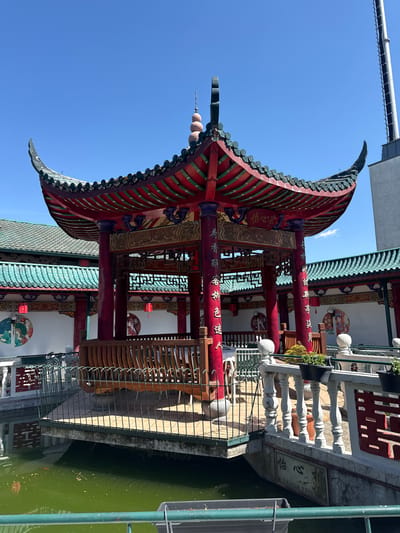 An outdoor, eye-level shot captures a vibrant, traditional East Asian-style pavilion, likely located at the Hainburg an der Donau location provided. The structure features a distinctive curved, multi-tiered roof adorned with red and green/blue tiles, topped with an ornate finial. Below the roof, rich red pillars stand prominently, embellished with intricate golden Chinese characters. Intricate wooden lattice work adds detail to the structure.
The pavilion overlooks a body of water, possibly a pond or canal, which appears green. A white balustrade with ornate spindles separates the pavilion's edge from the water. Potted plants are visible along the balustrade. The background reveals white walls adorned with circular decorative elements featuring traditional Chinese art motifs, such as figures in traditional dress and landscapes. The sky is clear blue, suggesting a sunny day. No people are visible, indicating a serene and tranquil setting.
Visible text on the pillars and a stone plaque includes:
* "怡心亭" (Yí Xīn Tíng), which translates to "Joyful Heart Pavilion."
* "荷香醉客新色迷人," a poetic phrase meaning "Lotus fragrance intoxicates guests, new colors are charming."
* "香" (Xiāng), meaning "Fragrance" or "Incense."
* "吃喝" (Chī Hē), meaning "Eat and Drink."
* "公元2002年9月8日" (September 8, 2002), indicating the date.