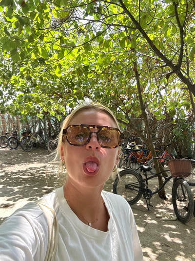A selfie depicts a woman with blonde hair wearing tortoiseshell sunglasses and a white shirt. She is sticking her tongue out towards the camera. Behind her, a sandy path is visible along with parked bicycles. Green foliage from trees creates a canopy overhead, casting shadows on the ground.