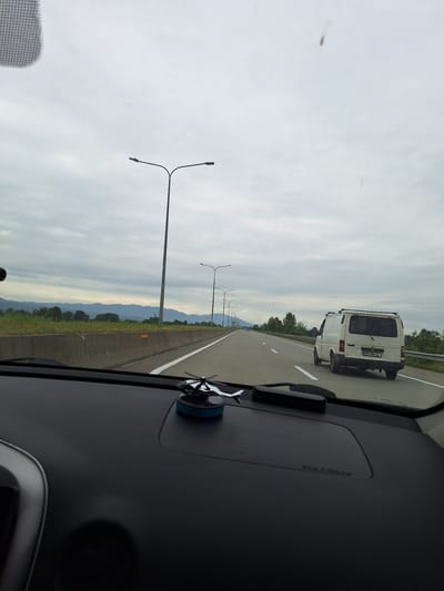 The image, taken from inside a vehicle, shows a highway scene under an overcast sky. The perspective is from the driver's seat, looking forward through the windshield. The highway has multiple lanes, marked with white lines. On the right side of the road, a white van with a license plate is visible, moving in the same direction. Streetlights line the side of the highway. In the distance, mountains and trees can be seen. The dashboard of the vehicle is in the foreground, with an air freshener and other accessories. The scene appears to be on a highway in or near Samtredia, Georgia, given the context.