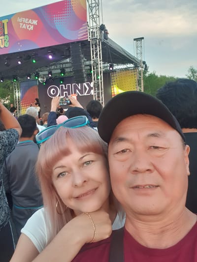 The image is a selfie taken outdoors at a public event, likely a festival or concert, in Taldykorgan, Kazakhstan. In the foreground, a man wearing a black baseball cap and a maroon t-shirt is on the right, and a woman with pink hair and large hoop earrings is on the left. She is wearing a white t-shirt and a maroon top, and has turquoise sunglasses perched on her head. Behind them, a stage is set up with a large screen displaying the text "OHNX" in white letters against a dark background. Above the stage, a colorful banner reads "ІДГЕАЖ ТАУКІ" in Cyrillic script. The sky is overcast. Numerous people are visible in the background, suggesting a crowd attending the event. The overall scene depicts a lively outdoor gathering in Taldykorgan.