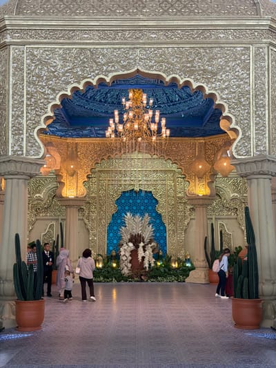 The image depicts the interior of a highly ornate building, likely a public space due to the presence of visitors. The architecture is characterized by extensive gold filigree and intricate designs, with large arched doorways and a prominent chandelier. A section of the building is colored a deep blue, adding contrast to the predominantly gold and beige color scheme.
Several individuals are visible within the building: one woman is dressed casually in a light-colored shirt and dark pants, carrying a pink bag, and there is a group consisting of a man in a suit, a woman in a patterned dress, and a child, suggesting a family outing. The individuals seem to be admiring the building’s design.
Tall cacti in large terracotta pots are placed as decorative elements, bordering the main walkway. The floor is a patterned carpet or tiled surface that complements the overall aesthetic of the interior.
Without additional reference, it is not possible to verify whether this location is accurately placed in the city of Pajang, Indonesia.