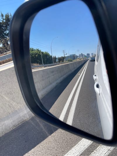 The image shows a reflection in a car's side mirror, depicting a highway scene near the city of Batzra, Israel. A white van is visible on the right side of the frame, occupying the immediate foreground of the reflection. The highway stretches forward, with clear lane markings consisting of double white lines. On the left side of the highway, a concrete barrier separates the lanes from the opposing direction. Beyond the barrier, trees and vegetation line the roadside, and in the distance, a skyline of modern buildings is visible, indicating an urban or suburban area. The sky is clear and blue. There are no people visible in the reflection. The overall scene suggests a typical daytime view from a moving vehicle on a highway.