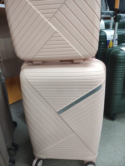 The image shows a close-up of stacked luggage, primarily a light pink suitcase on top of another similar suitcase. The luggage has a ribbed texture and a geometric pattern. To the right of the pink luggage, a dark green suitcase is visible. The floor appears to be carpeted. The scene is indoors, likely in a retail setting. There is no visible indication of the city of Oral, Kazakhstan within the image itself or the provided crops.