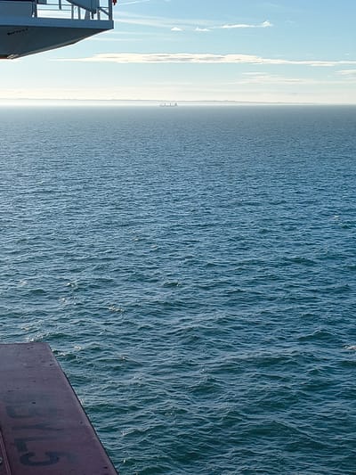 This media file features a vertical shot of a vast expanse of water, likely the sea, with a hazy sky in the background. On the left side of the image, a portion of a metallic structure, possibly part of a ship or a dock, is visible. The structure has white and grey elements, including railings and a slanted surface. The water is a deep blue and appears somewhat choppy, with light reflecting off its surface. The sky is a pale blue with streaks of white clouds, creating a sense of distance and possibly indicating a slightly overcast or hazy day. The horizon line is barely discernible due to the haze.
In the far distance, on the horizon where the water meets the sky, there are faint outlines of what might be structures, possibly a coastline or offshore installations. The overall mood is one of openness and vastness, with the natural elements of water and sky dominating the scene. The light suggests it could be daytime, perhaps morning or late afternoon, given the soft quality of the light.
Visible text includes "3YL5" in large, faded, dark grey letters on the metallic structure on the right side of the image. The image's orientation is vertical, with the water occupying the majority of the frame.