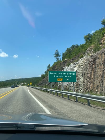 The image depicts a highway scene on a sunny day. A green highway sign indicates "Grenville-sur-la-Rouge Chemin Avoca" with a route number "226" and an arrow pointing to the right. The sign is positioned on the right side of a multi-lane highway. The highway runs through a landscape with rocky cliffs and lush green trees on the right, and a forested hillside on the left. Several cars are visible on the road. The foreground shows the hood and windshield of a vehicle, suggesting the photo was taken from a car. The sky is blue with some scattered clouds. The location is identified as Pointe-au-Chêne, Canada.