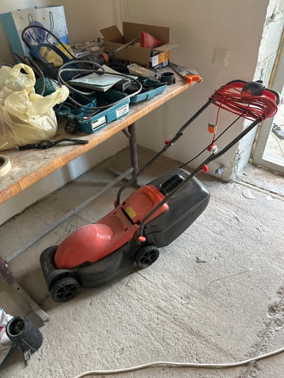 In an indoor setting with a concrete floor and unfinished walls, a red and black electric lawnmower is the prominent object. It is positioned diagonally on the floor, with its handle and tangled orange electrical cord extending upwards and to the left. The lawnmower's grass box is black, and the main body is a bright red. The "Flymo" brand name is visible on the lawnmower's casing.
To the left of the lawnmower, a wooden shelf is laden with various tools and supplies. A blue and white patterned gift bag is at the bottom of the shelf, and on top of it, a yellow plastic bag contains what appears to be gardening supplies. Tools such as a blue drill and a green electric device are also present. A black digital watch is laid out on the shelf, next to a roll of tape. In the background, cardboard boxes are stacked, and a red funnel is visible. The scene suggests a storage area or a workshop, possibly during home renovations given the unfinished concrete and walls. The lighting indicates it is daytime, with natural light filtering in from an unseen source. No people are visible in the image.