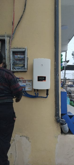 An outdoor wall installation in Aktash, Uzbekistan, is depicted forensically. The light beige wall, showing minor cracking and discoloration at its base, features mounted electrical equipment. A person, viewed from behind and positioned on the left, wears a dark plaid long-sleeved shirt and dark trousers, with their hands near the electrical components. An open metal electrical box containing multiple circuit breakers and wiring is affixed to the wall above a smaller, open electrical panel also housing circuit breakers. To the right of these panels, a white rectangular power inverter is mounted, displaying "kW" and "ON" in orange text on its digital screen. Three blue conduits connect the smaller electrical panel to the bottom left of the inverter, while two black cables connect to its bottom right. Additional black and grey electrical cables run vertically along the wall from the top of the frame. A dark grey vertical drainpipe is visible along the right edge of the frame, terminating in a grey PVC elbow near the bottom. The blurred background on the right reveals blue structures, stacked green cylindrical objects, and an overcast sky, consistent with an outdoor commercial or industrial environment.