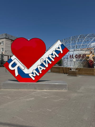 An outdoor, low-angle, medium shot captures a large, stylized red heart sculpture in the foreground, featuring the Russian flag's white, blue, and red stripes. The words "Я МАЙМУ" (I Love Mayma) are prominently displayed on the sculpture in white, illuminated letters. The sculpture is situated on a metallic platform in a paved public area, likely a town square. In the background, a temporary stage structure with white metal supports is visible, adorned with a large banner that appears to advertise an event. The banner features Cyrillic text, an image of a person in traditional attire, and QR codes. A brick building with a modern facade and a few evergreen trees are also present in the left background, under a clear blue sky. The scene suggests a public gathering or event in the town of Mayma, Russia.