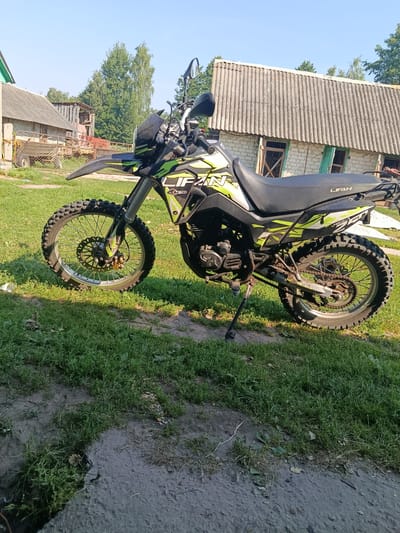 A dirt road passes through a grassy field with a motorcycle parked on the grass. The motorcycle is a green and black dirt bike. In the background, there is a dilapidated wooden building with a corrugated metal roof and a brick building with a similar roof. There are trees and a clear blue sky in the background. The scene is likely in a rural area of Khynochi, Ukraine, based on the architecture and vegetation.
