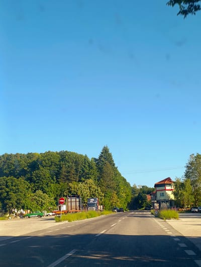The image depicts a rural road scene in Panichkovo, Bulgaria, on a clear, sunny day. The road, paved and marked with white lines, curves slightly towards the right. On the left side of the road, there is a parking area or lot with several parked vehicles, including a vintage green car and a dark-colored truck with a trailer. A circular red and white "no entry" sign is prominently displayed near the parked vehicles. Beyond the parking area, a dense line of lush green trees forms a backdrop.

On the right side of the road, a two-story building with a red-tiled roof stands, featuring a green and yellow facade. A "30" speed limit sign is visible near this building. More trees and foliage line the right side of the road, extending into the distance. Several other vehicles, including cars and a utility vehicle, are present on the right side of the road and in the distance, suggesting a small settlement or business area. The sky is a clear, bright blue with a few faint circular smudges, likely from lens imperfections. The overall impression is one of a tranquil, sunlit rural environment with signs of moderate activity.
