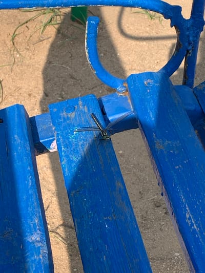 A close-up shot captures a dragonfly perched on a weathered, bright blue wooden bench. The bench is part of outdoor furniture, with visible signs of wear and tear on the paint. The background consists of sandy ground with sparse blades of dry grass and the shadow of what appears to be the bench. The dragonfly is in focus, displaying its intricate wings and segmented body. The scene suggests a potentially rustic or natural setting within the city of Тавричанка, Russia, where outdoor seating is available.