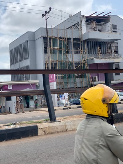 The image shows a partially collapsed building under construction in the city of Ondo, Nigeria, located at coordinates 7.252 latitude, 5.199 longitude. A large, gray building is partially wrapped in scaffolding made from bamboo poles. Part of the roof has collapsed and is now lying on the ground. There are people walking on the street in front of the building, including a man wearing a yellow helmet. The street is paved and has black and white curbs. A sign for "Wema Bank" is visible on the building. There is an "ATM 24/7" sign on a smaller, pink building to the left of the main structure. A black metal railing blocks most of the image from view. The sky is cloudy and there are power lines running across the top of the building.