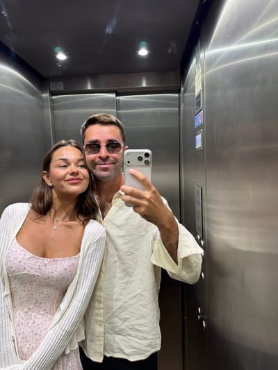 A couple is taking a selfie in an elevator. The man, on the right, is wearing a light-colored button-down shirt and sunglasses. He is holding a phone to take the photo. The woman, on the left, is wearing a light-colored cardigan over a floral-patterned dress and a necklace. They are standing close together, with the woman leaning her head on the man's shoulder. The elevator has brushed metal walls and ceiling lights. The image has a mirror-like quality, reflecting the interior of the elevator. The specific location in Pognana Lario, Italy, is not discernible from the image itself.