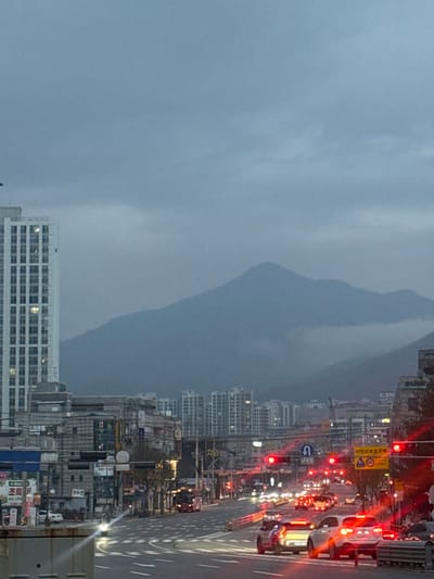 A cityscape with a mountain backdrop dominates the image. The sky is overcast, casting a muted light over the scene. A busy road with traffic lights and vehicles runs parallel to tall buildings. The architecture is a mix of residential and commercial structures. The mountain appears as a soft, blurry shape in the distance. The image is rotated counterclockwise by 90 degrees.
