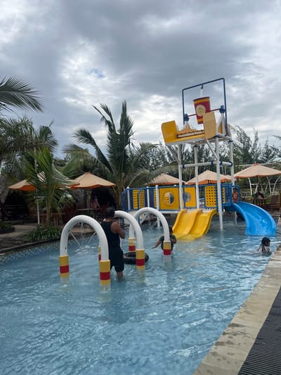 This image depicts an outdoor water park facility located in Juwana, Indonesia, under an overcast sky. The foreground and mid-ground are dominated by a blue-tiled swimming pool. In the shallow end, three white, arch-shaped water sprinklers with red and yellow bases are actively spraying water. An adult male in a black tank top and shorts stands among these sprinklers, with his left foot on a black inner tube. A small child in a green top stands nearby, also in the shallow water. Further right in the pool, another child, partially submerged, is visible near the edge.
Central to the scene is a multi-tiered water play structure, predominantly yellow and blue, featuring two yellow water slides and one blue curved water slide descending into the pool. At the top of this structure, a large red and yellow tipping bucket is suspended. The background displays lush tropical vegetation, including numerous palm trees and other dense green foliage. Several thatched-roof bungalows or huts, equipped with tables and chairs, are visible to the left and in the far background, some with indistinct figures seated. Orange patio umbrellas are also scattered throughout these resting areas. The overall environment suggests a recreational leisure setting.