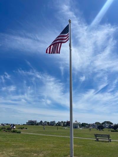 An American flag flutters against a bright blue sky filled with wispy white clouds. The flag is attached to a tall flagpole that extends horizontally across the frame. In the background, a well-maintained green lawn stretches out, lined by a gravel path. Beyond the lawn, a row of charming houses with varied architectural styles sits nestled amongst trees. Further in the distance, more houses and buildings are visible, suggesting a residential area. A few people are scattered across the lawn, some walking and others appearing to be relaxing. The scene evokes a sense of a pleasant day in a seaside town, likely in the United States, given the prominent display of the American flag. The lighting suggests it is daytime, possibly late morning or early afternoon, with clear and fair weather. There is no visible text in the image. The overall mood is peaceful and idyllic.