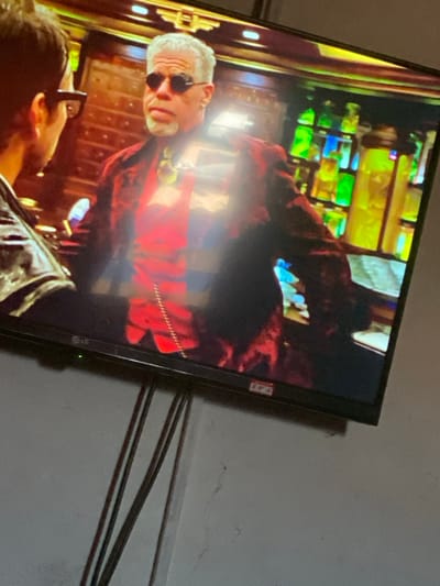 This image depicts a television screen displaying a scene from a film or series, captured within a room in Mape, Nigeria. On the screen, a fair-skinned male with grey hair and beard, sporting small dark round sunglasses, a red patterned blazer over a red shirt, and a yellow tie with a prominent green medallion, is shown from the chest up. A silver chain hangs from his left jacket pocket. He faces another individual, seen partially from the back, wearing dark-framed glasses and a dark jacket. The background of the television scene features dark wood paneling and shelves filled with illuminated glass vials or bottles in vibrant green, yellow, and blue hues. The television itself is a black-framed flat-screen LG model, featuring a green indicator light on the bottom left. Multiple dark cables descend from behind the television against a light grey wall that exhibits minor stains or discoloration.