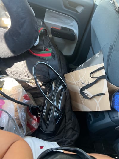 The image shows the interior of a vehicle, likely parked. The scene is cluttered with various personal items and bags. In the foreground, part of a person's legs and lower torso are visible, wearing white underwear with a small red heart design.
Dominating the center of the frame is a black, patent leather handbag with a shiny finish and silver hardware. To the left of this handbag, a larger black quilted bag with a Gucci logo pattern is partially visible. A red and green striped ribbon is draped across it. Also on the left, a black neck pillow is positioned near the top of the frame.
In the background, a light brown paper shopping bag with black ribbon handles is visible. The bag has a design with intertwined white lines and text that appears to read "SHOP HOUSE". A blue item is peeking out from behind this bag. The seats and door panel of the vehicle are visible, indicating it is a car interior. The lighting suggests it is daytime, with sunlight illuminating parts of the scene.
The provided metadata indicates the location is North Charleston, United States of America.