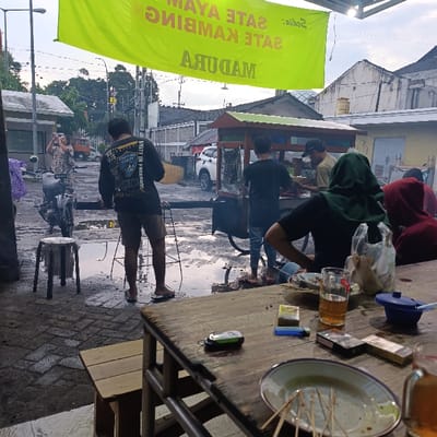 A group of people are gathered outside a food stall in Kecamatan Asembagus, Indonesia. The stall is located at -7.749 latitude and 114.218 longitude. The stall is a small, open-air structure with a metal roof. There are two people inside, preparing food. One person is standing and the other is seated. A woman wearing a blue shirt is standing near a motorcycle. The man in the front wearing a black shirt and blue shorts is facing the stall, while another man seated in the foreground is looking towards the camera. A man in a green shirt and a man in a black shirt are standing near the stall. The street is wet, and there is a small stool near the motorcycle. A wooden table with multiple items on it is in the foreground. There is a bowl with chopsticks, a glass of liquid, cigarette packs, and a lighter on the table. The scene is captured during the day, with the sunlight illuminating the area.