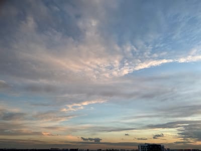 The image depicts a panoramic view of the sky over an urban landscape, likely at sunset or sunrise. The sky is filled with a variety of clouds, ranging from wispy, streaky formations to denser, more textured masses. The colors in the sky transition from a pale blue at the top to softer hues of pink, orange, and yellow near the horizon, indicating the presence of the sun just below the visible frame.
In the lower portion of the image, a city skyline is visible. Silhouettes of buildings and structures are discernible against the lighter sky. Some buildings appear to be of modern design, with multiple stories and visible windows. Construction cranes are also present, suggesting ongoing development or the presence of a construction site. The overall impression is one of a city at the edge of day, with the natural beauty of the sky contrasting with the man-made environment. The image is taken from an elevated vantage point, offering a broad perspective of the scene.