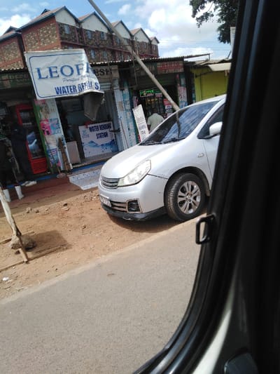 The image displays an outdoor street scene in Kikuyu Township ward, Kenya, captured from the interior of a vehicle looking through its passenger-side window. In the foreground, an asphalt road is visible, adjacent to an unpaved dirt shoulder that runs parallel to the road. A silver passenger vehicle, identified by its front bumper, grille, headlights, and front passenger wheel, is positioned on this dirt shoulder, facing towards the right. Its lower front bumper/spoiler is black.

Along the dirt shoulder, a series of single-story commercial structures are present. Most prominently, a shop in the mid-left features a large blue and white sign reading "LEOFRESH Purified Drinking WATER REFILL STATION." Inside this establishment, a secondary sign stating "WATER VENDING STATION" is visible, along with a red beverage cooler positioned to the far left of the entrance. A person in dark attire and white footwear stands partially visible near this cooler, with their back to the viewer. Immediately to the right of the LEOFRESH shop, another shop with a corrugated metal roof displays a green M-PESA sign; a second individual, wearing a light-colored shirt, is visible inside this shop, facing away from the viewer. Further right, a yellow-painted shop with a similar roof structure is partially discernible.

In the background, a multi-story building featuring a red and brown brick-patterned facade and multiple white-trimmed gabled roof sections rises above the commercial strip. A light-colored pole extends diagonally across the upper portion of the scene. The sky above is partly cloudy, and the entire environment is illuminated by natural daylight.