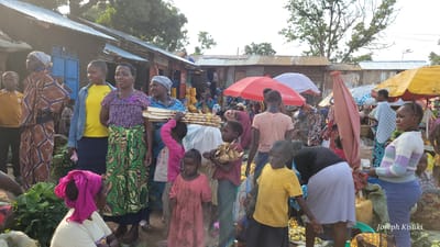 The image depicts a bustling outdoor market scene in Bunia, Democratic Republic of the Congo. Multiple individuals, primarily women and children, are present. The foreground shows several people, including a woman in a pink headscarf and yellow shirt, a young boy in a pink shirt carrying wooden sticks, and a girl in a pink dress. To their left, a woman wearing a distinctive blue and brown patterned dress stands with her back to the viewer, while another woman in a yellow shirt and blue skirt is visible. In the center, a woman in a purple floral top and green patterned skirt carries a stack of wooden items on her head. Several market stalls are visible in the background, constructed with corrugated metal and wooden planks, some covered by colorful umbrellas, including a prominent red one. Various goods, possibly produce and other merchandise, are displayed on the ground and in stalls. The atmosphere appears lively and crowded, typical of a local market.