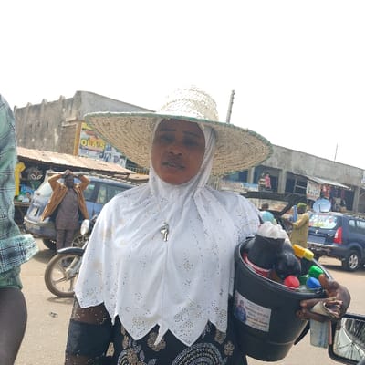 The image depicts an urban street scene in Olomoda, Nigeria. In the foreground, a woman faces forward, wearing a broad, light-colored woven straw hat over a white hijab and a dark, patterned garment. A silver-colored pendant hangs from her neck. She holds a black plastic bucket in her left hand, which contains multiple bottled beverages with colorful caps (red, green, yellow, blue) and some with dark liquid. A sticker with text and a man's portrait is affixed to the bucket. Her right hand, partially visible, holds folded currency.
In the mid-ground to the left, a man in a brown jacket and dark trousers stands with both hands on his head, positioned near a parked blue van and the front wheel of a motorcycle. The background features several multi-story concrete buildings, some with visible shopfronts and merchandise. A yellow and red sign bearing the text "OLA'S" is visible on one building. To the right, a blue car is parked, and an additional figure is partially visible further back. The ground appears to be a paved road, and the sky is overcast or brightly lit.