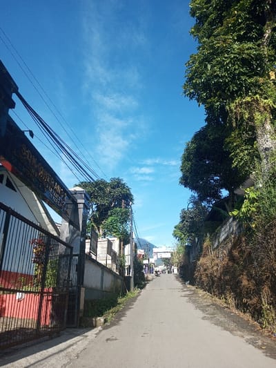 The scene depicts a paved street ascending an incline under a clear blue sky with sparse cirrus clouds in Khouathineung, Laos. On the left, a dark metal gate, partially open, reveals a white building with red accents. An overhead sign on the gate structure displays text including "SDN CIKERANG 2", "JL. KARET NO. 1", and "CIKERANG, KAB. BEKASI". Multiple black electrical wires traverse the upper left of the frame, originating from a utility pole near the gate. A concrete wall runs alongside the left of the road, bordered by green vegetation. On the right, a large tree with dense green foliage and climbing vines dominates the foreground, with dry, brown shrubbery and another concrete wall running alongside the road. The street, which lacks discernible markings, leads uphill towards a distant archway structure. Several motorcycles and indistinct figures are present further up the road. A mountain or large hill is visible in the distant background under the bright sunlight.