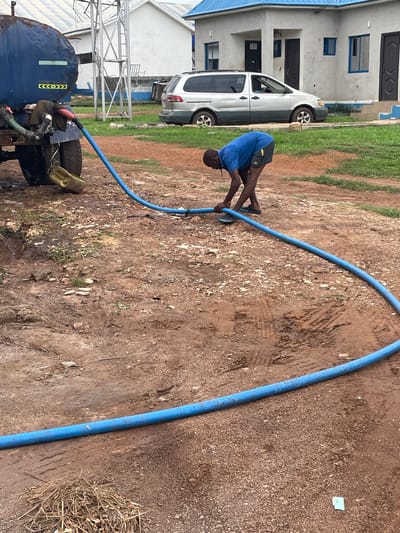 A person is seen bending over while holding a blue hose in an outdoor scene. The person is wearing a blue shirt and dark shorts. The setting appears to be a dirt ground with some patchy grass and scattered small stones. A white vehicle and a building with blue trim are visible in the background. There is also a large dark blue tank on a trailer with tires, suggesting it might be a water truck. The hose is connected to the tank and extends towards the person. The overall impression is that the person is operating or managing the hose, possibly for filling or emptying the tank. The lighting suggests it is daytime. There are no discernible emotions visible. There is some text on the side of the tank, which appears to be yellow and black hazard symbols.