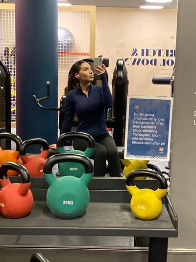 A woman is taking a selfie in a gym. She is wearing a dark blue long-sleeved shirt and black leggings. She has dark hair and is wearing earbuds. Behind her, various kettlebells are neatly arranged on a rack, with weights ranging from 8 KG to 30 KG. The kettlebells are red, green, and yellow. A sign with Swedish text is displayed on the wall, informing gym-goers about the proper use and return of the heavier dumbbells. The background includes gym equipment and a blue wall. The overall setting appears to be a modern, well-equipped fitness facility. The image is oriented vertically, suggesting it was taken with a phone. The lighting indicates it is daytime indoors.