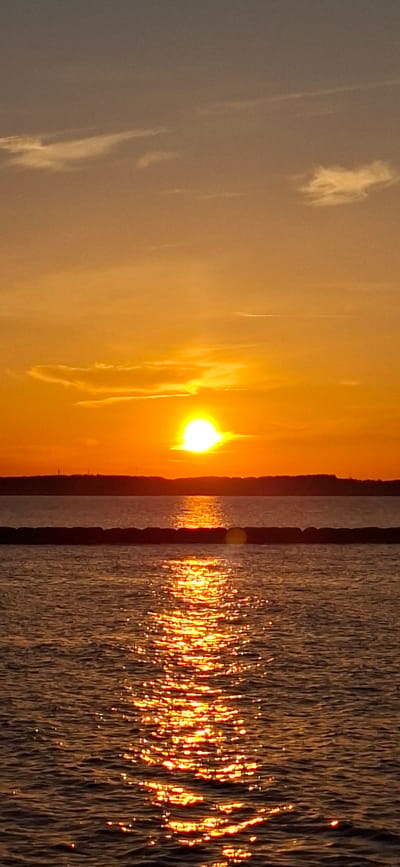 A vertical, low-angle shot captures a sunset over the water in Pärnu linn, Estonia. The sky is a vibrant gradient of orange and yellow, with wispy white clouds scattered across the upper portion. The sun, a bright, glowing orb, is positioned slightly left of the center, casting a brilliant reflection on the water. A dark, low horizon line separates the sky from the body of water. In the middle ground, a dark, segmented object, possibly a breakwater or a series of buoys, stretches horizontally across the image, partially submerged in the water. The foreground is dominated by the rippling surface of the water, which shimmers with the reflection of the sunset, creating a pathway of golden light. The overall scene conveys a sense of tranquility and natural beauty.