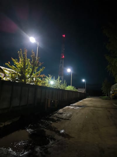 The image depicts a rural nighttime scene, possibly in the vicinity of Бабініцкі сельскі Савет, Belarus. A dirt road, wet and reflecting the light, stretches into the darkness. Puddles of water are visible on the road. A tall, red and white lattice tower, likely a telecommunications tower, is illuminated with red lights at its top. Streetlights are present, one casting a bright glare on a tree with green and orange foliage, and another further down the road illuminating the path. A concrete fence runs along the left side of the road, partially obscured by trees and a building in the background. The environment appears to be a quiet, possibly industrial or residential area on the outskirts of a settlement. There are no people or animals visible in the image.