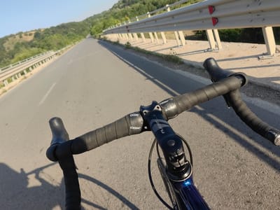 The media file shows a first-person perspective of someone riding a bicycle on an asphalt road. The handlebars of the bicycle are prominent in the foreground, wrapped in black tape and branded with "COX". The bicycle's stem is also visible, with the word "FLIGHT" printed on it. A dark blue bicycle frame is partially visible below the handlebars.
The road stretches ahead, curving slightly to the left, with a guardrail on the right side. Beyond the guardrail, the landscape consists of rolling hills covered in green vegetation and trees, indicating a rural or semi-rural setting. The sky is clear and blue, suggesting fair weather. The lighting indicates it is likely late afternoon or early morning, with long shadows cast by the guardrail and potentially the cyclist. The location is identified as Staliyska mahala, Bulgaria. There are no people visible in the frame. The overall impression is one of a peaceful cycling journey through a scenic, hilly landscape.