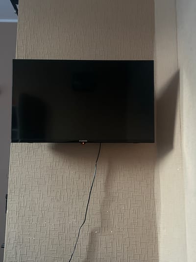 A flat-screen television is mounted on a textured, beige wallpapered wall. The TV is black and appears to be off, with a small, illuminated red indicator light visible on the bottom right corner of its frame. A black cable hangs down from the television, extending towards the bottom of the frame. To the right of the television, a shadow is cast on the wall, indicating a light source from the left. The environment appears to be indoors, likely a room in a residential building. The image does not provide enough information to determine the specific location within Давыдовка, Ukraine, or any details about people or their actions.