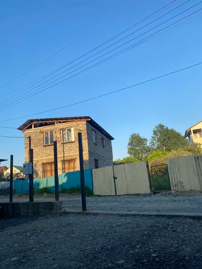 This image depicts a residential scene in Verkhnevesyoloye, Russia, under a clear, bright blue sky with direct sunlight suggesting either early morning or late afternoon, as evidenced by long shadows cast to the left. In the midground, a two-story house constructed of grey cinder blocks dominates the left-center. The house appears to be either unfinished or in disrepair, featuring a partial, rusted metal roof with exposed wooden rafters and debris visible beneath. The upper floor has two windows, one with a vertical blind and the other with a simple frame. The lower floor windows are obscured by decorative metal security grates, behind which dark coverings are visible.

In the immediate foreground, a gravel and dirt surface constitutes an unpaved area. Several dark, rusted metal poles are vertically embedded in the ground, one of which supports a grey electrical utility box on its left side. Behind these poles and to the left of the main house is a turquoise corrugated metal fence, casting a prominent shadow. To the right of this fence, a light-colored wooden slat fence stands, with a section open to reveal a dark, ornate metal gate leading into a grassy area.

Beyond the main house and fences, a dense cluster of green trees is visible in the background, and to the far right, a portion of another house with a light-colored facade and a balcony can be seen. In the far left background, other residential structures with roofs are partially visible. Overhead, five parallel power lines stretch diagonally across the blue sky from the upper left to the lower right. No people or animals are present in the scene.