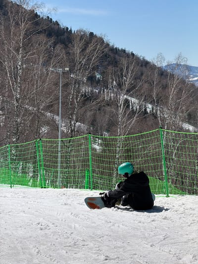 A snowboarder in black attire and a teal helmet is on a snowy slope next to a green safety net, possibly on a ski resort. The location is likely near Ozernoe, Russia, as specified. Behind the net and slope are bare trees against a light blue sky.