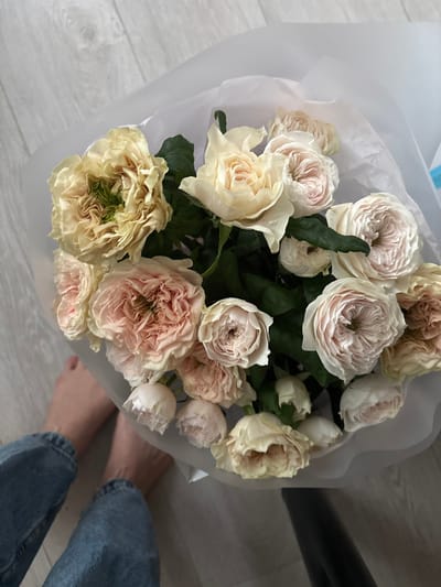 A close-up, overhead shot captures a bouquet of light-colored roses, predominantly shades of cream and blush pink, artfully arranged in white tissue paper. The roses exhibit a tightly packed, ruffled petal structure, with some blooms appearing more fully open than others, revealing hints of green at their centers. Dark green leaves are interspersed throughout the floral arrangement. In the lower left quadrant of the image, a pair of bare feet and the lower legs of a person wearing faded blue denim jeans are visible, suggesting they are standing or sitting on a light-colored wooden floor. The flooring exhibits a subtle grain pattern. The background consists of the same wooden floor and a plain white surface, likely part of the bouquet's wrapping or a nearby surface. There is no visible indication of the city of Сокол, Russia, within the image itself. The lighting appears soft and diffused, creating subtle shadows and highlighting the textures of the petals and the wood grain.