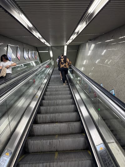 The image, taken in 龙溪街道, China, shows a perspective view from above of a set of escalators within what appears to be a subway or underground transportation hub. Two escalators are visible. One is operating and has two people riding it, while the other has one person lying on it. The surroundings include architectural details such as walls with linear patterns, lighting fixtures, and tiled flooring. There are posters or advertisements fixed on the walls.